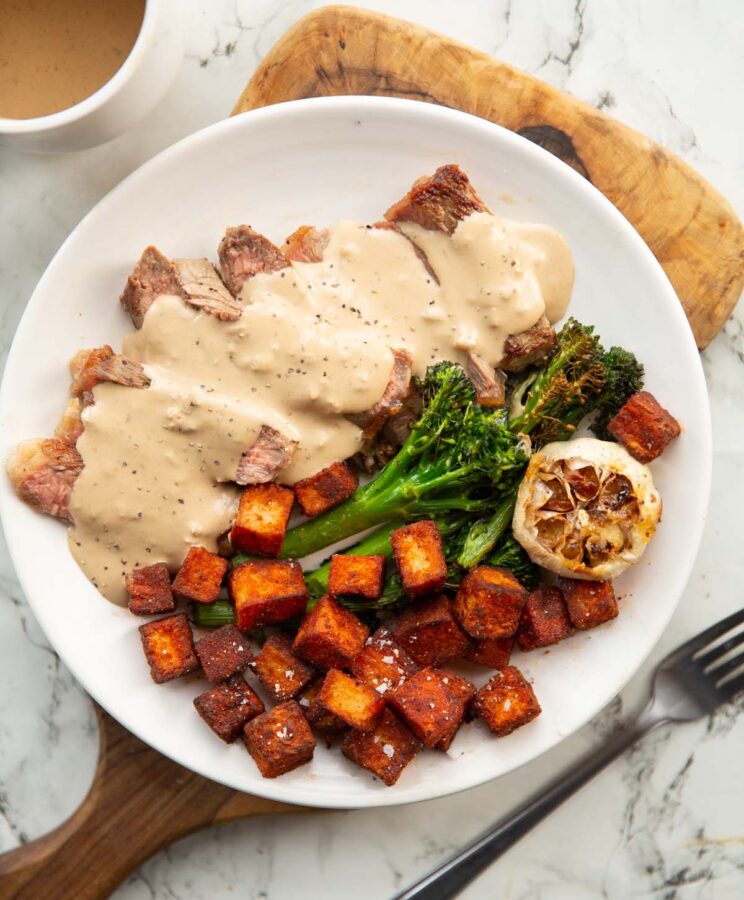 sliced steak with creamy garlic sauce on white plate with potatoes and broccolini