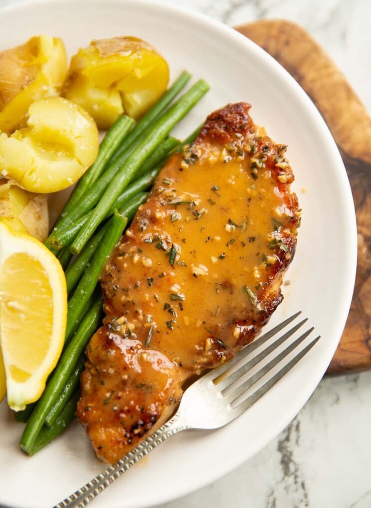 close up shot of garlic butter pork chop with green beans and baby potatoes on small white plate with silver fork