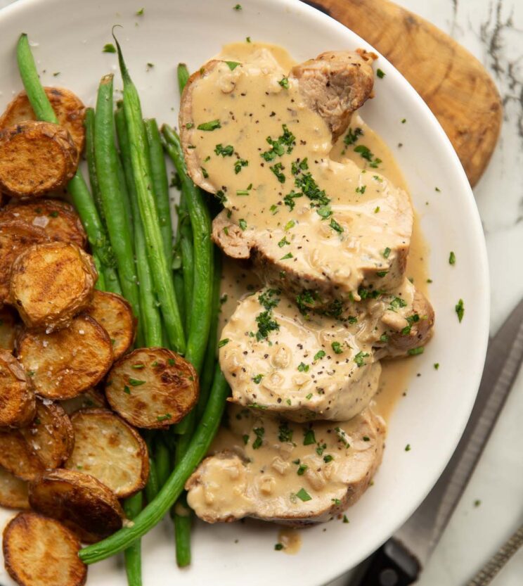 overhead shot of sliced pork tenderloin with boursin sauce on white plate with potatoes and green beans