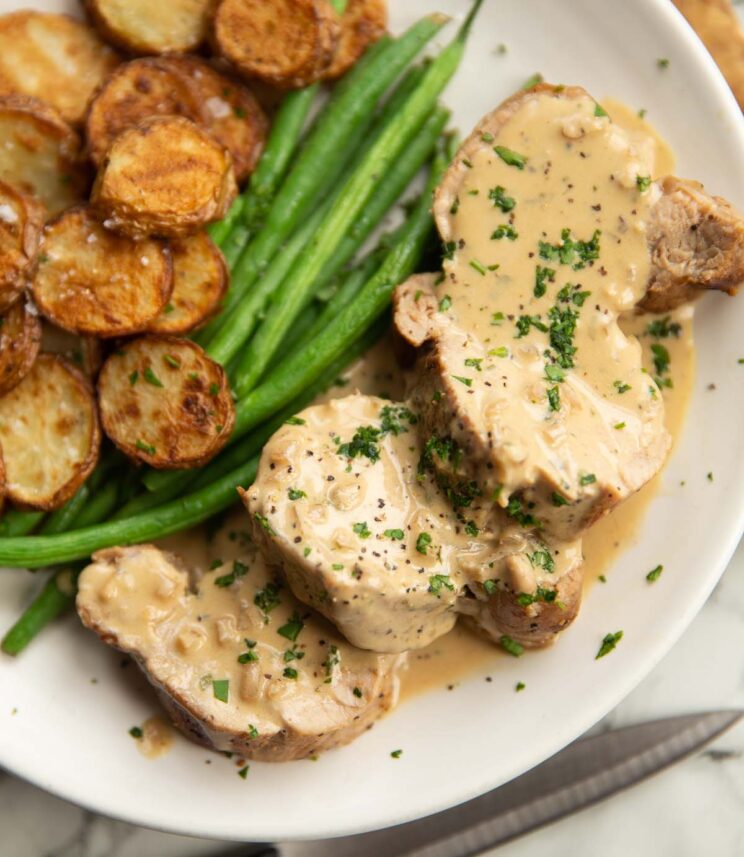 close up shot of sliced pork tenderloin with boursin sauce on white plate with green beans and potatoes