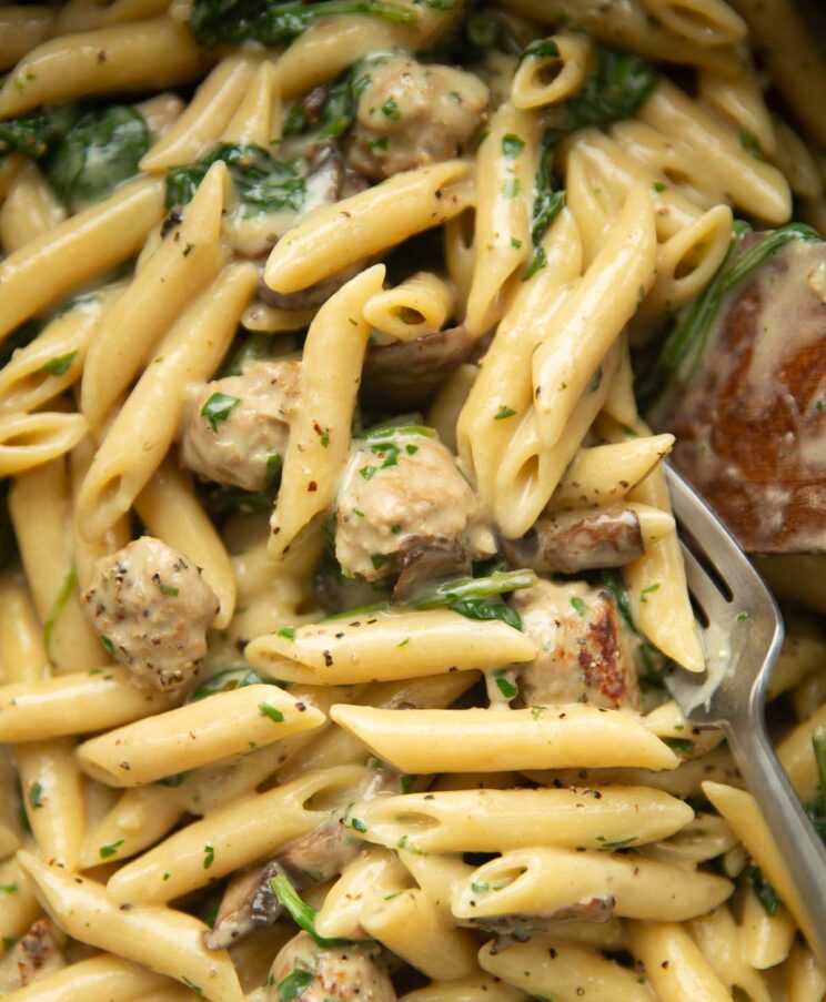 close up shot of mushroom and sausage pasta in large pot with fork and wooden spoon digging in