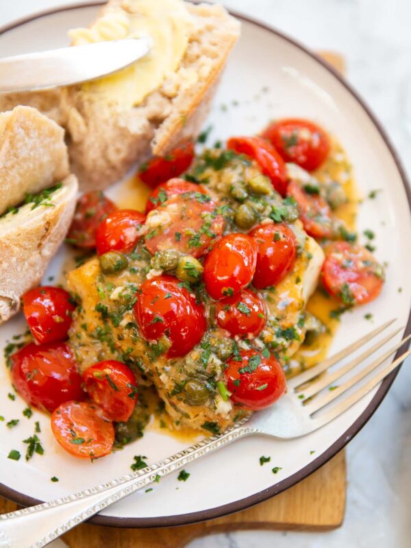 cod with tomatoes and capers served on small white plate with bread and silver fork