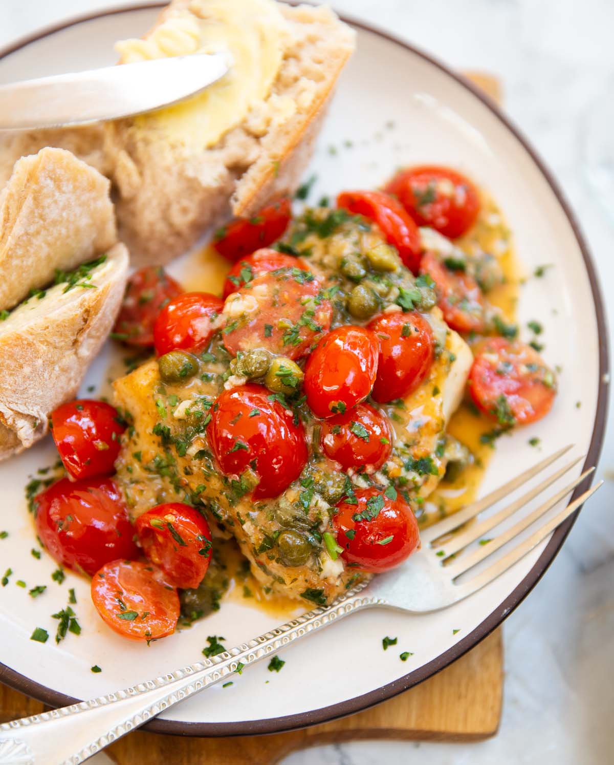cod with tomatoes and capers served on small white plate with bread and silver fork