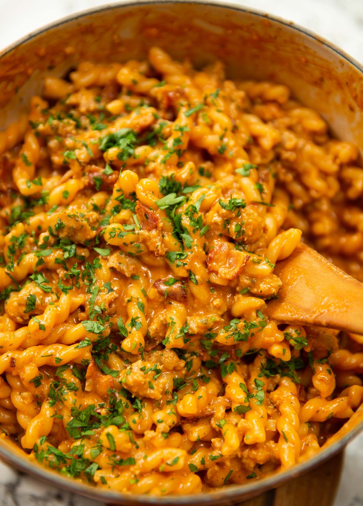 sausage and bacon pasta in large pot with wooden spoon garnished with fresh parsley