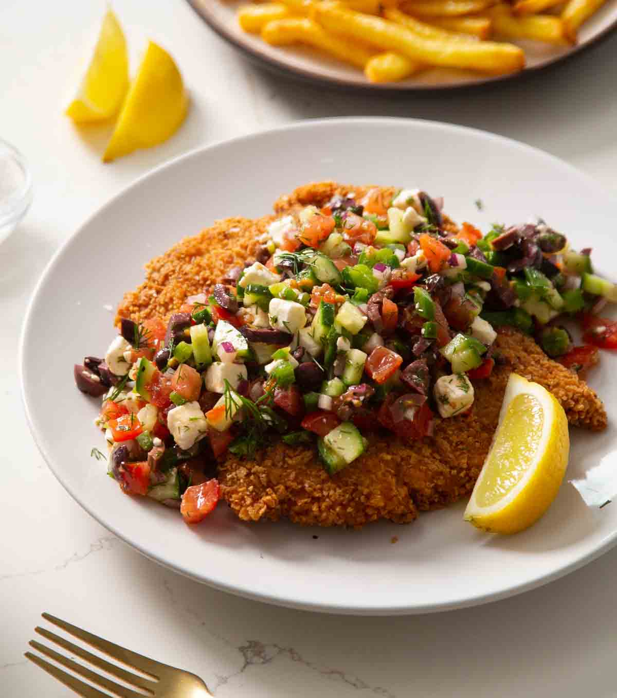 crispy greek chicken on a white plate with fries blurred in the background