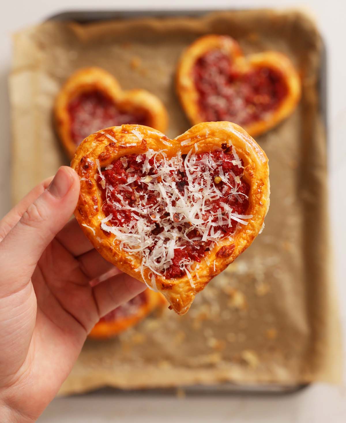 overhead shot of hand holding puff pastry pizza love hearts above baking tray