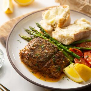 cajun baked cod on small white plate with asparagus, tomatoes and bread