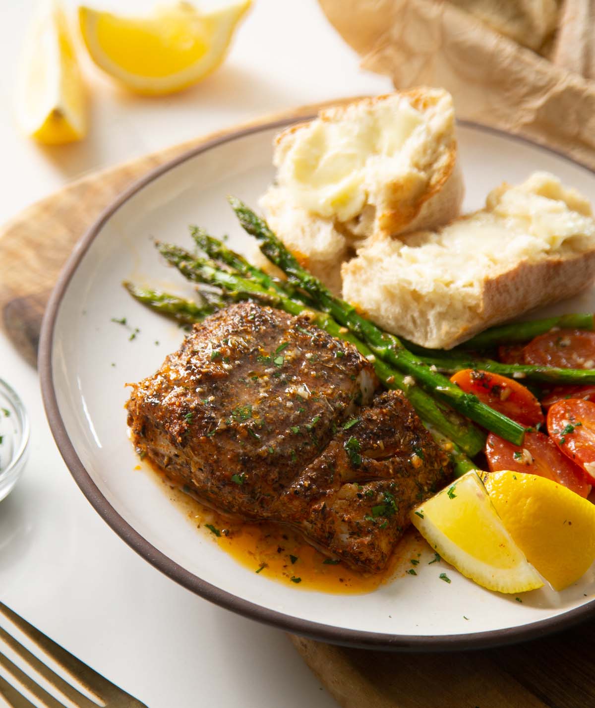 cajun baked cod on small white plate with asparagus, tomatoes and bread