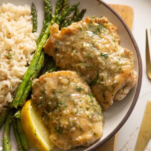 2 chicken thighs on small white plate with rice and asparagus and lemon wedge
