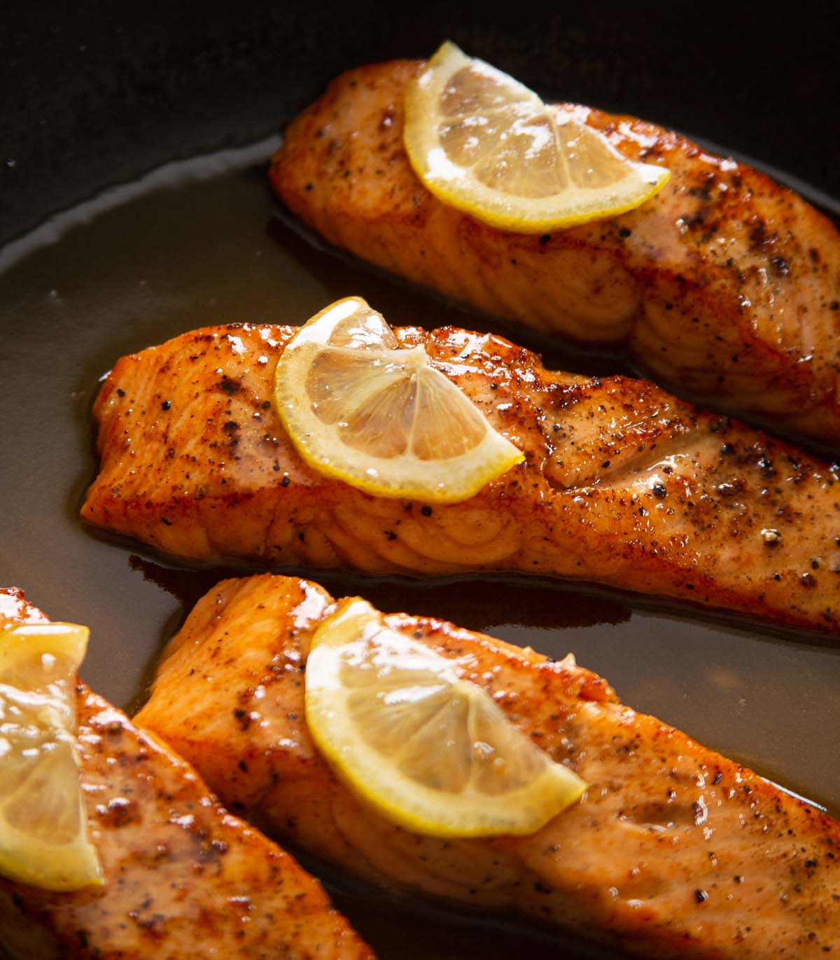 close up shot of 4 salmon fillets in pan garnished with lemon