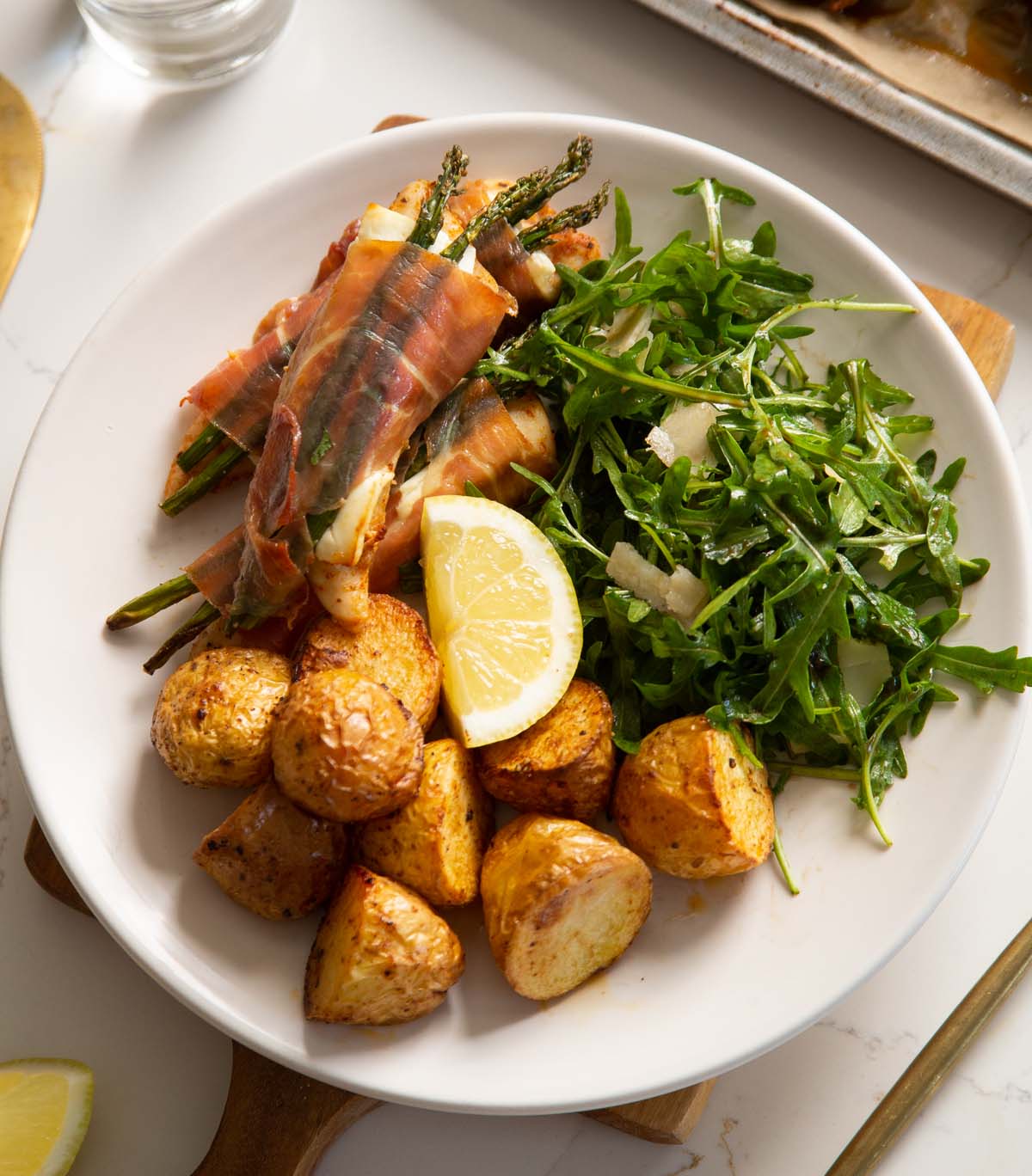 prosciutto wrapped chicken on small white plate with potatoes, salad and lemon wedge