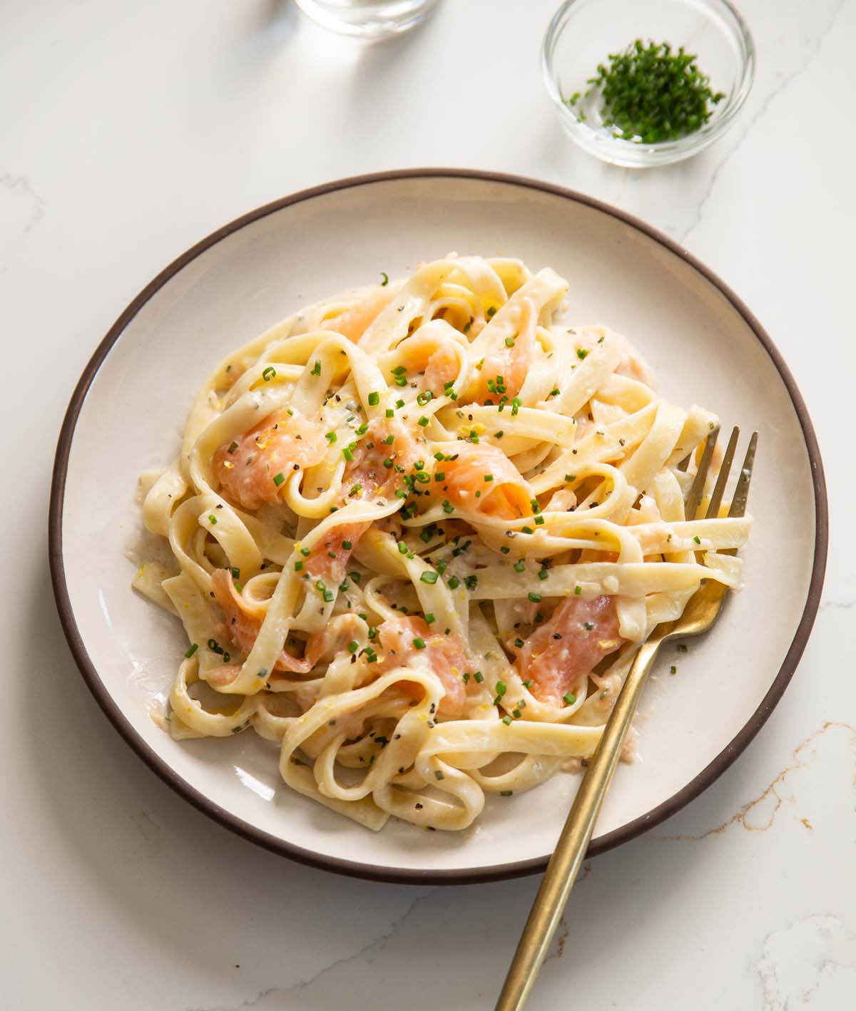 smoked salmon pasta served on small white plate with gold fork and small glass pot of chives in background