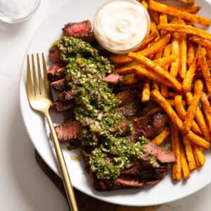 close up shot of steak and chimichurri served with fried on white plate with gold fork