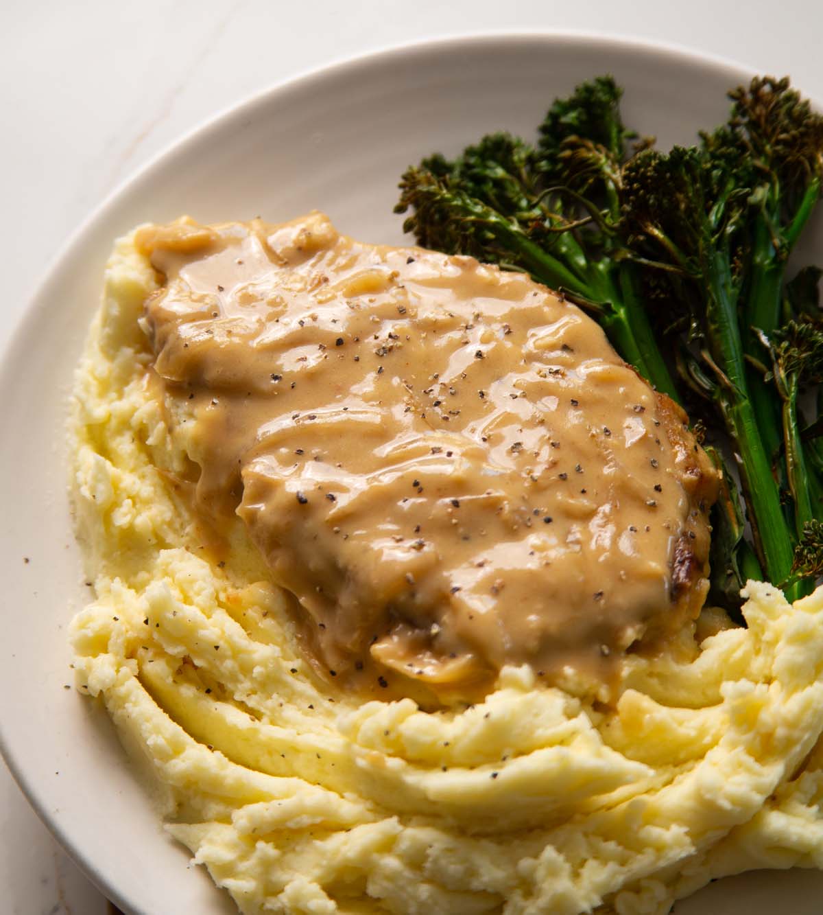 smothered pork chop served on small white plate with mash and broccolini